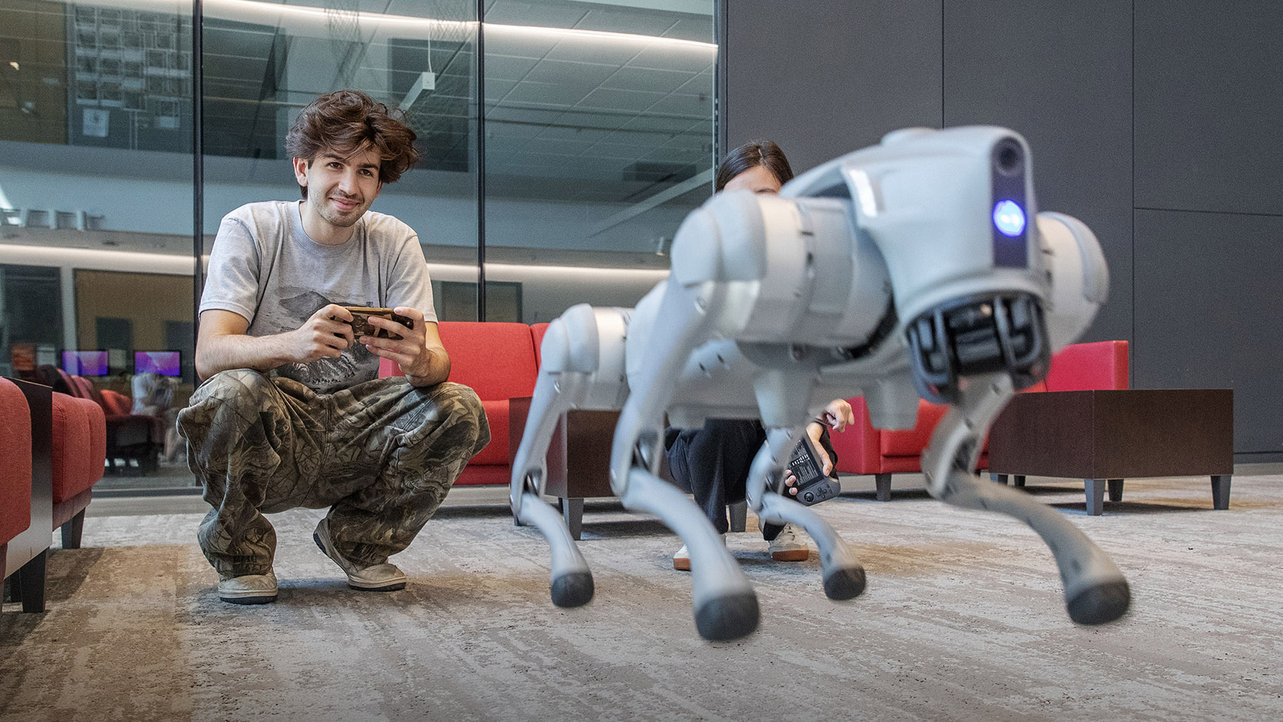 A student crouches with a handheld controller while a four-legged robot walks toward the camera in a campus building