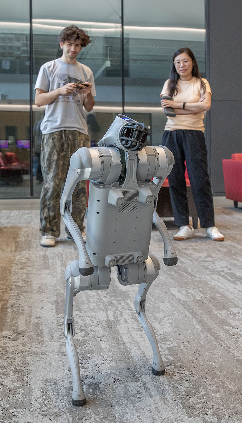 A four-legged robot standing in a campus building while a student operates it with a handheld controller