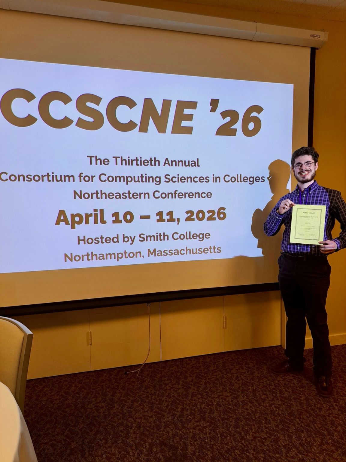 Josh Feldman holding his first prize certificate beside a screen reading CCSCNE &rsquo;26, the Thirtieth Annual Consortium for Computing Sciences in Colleges Northeastern Conference